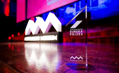 A photo of a stage, lit up with pink and blue lights, and a glass trophy in the foreground saying Eureka Prizes.